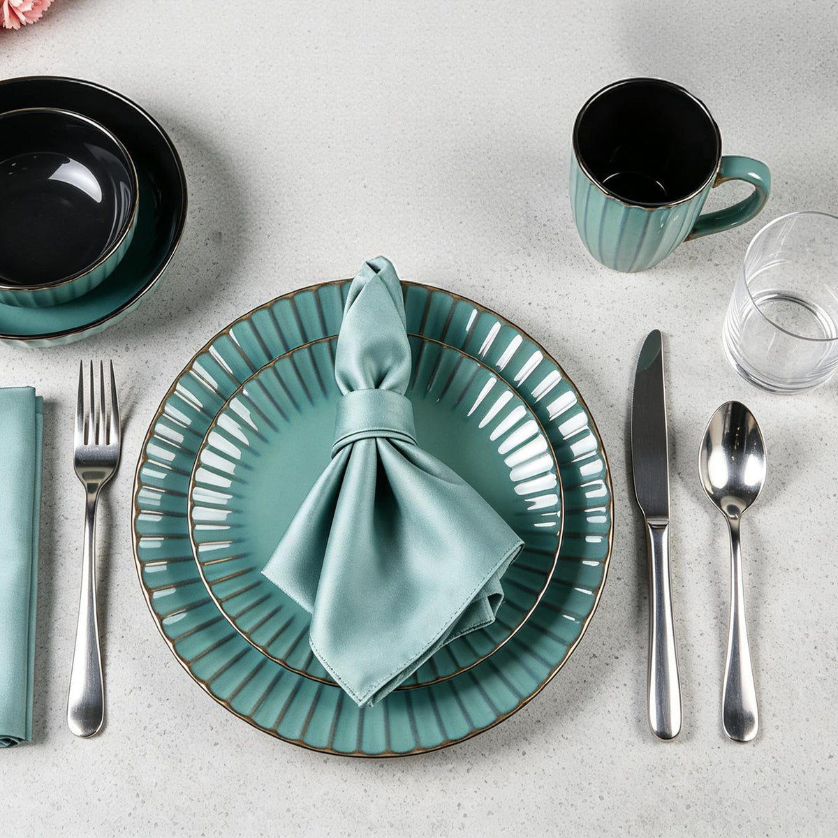 A styled table setting on a light stone surface featuring a teal ribbed ceramic dinner plate with a matching napkin, accompanied by silver flatware, a black and teal bowl, a matching mug, and a clear drinking glass.