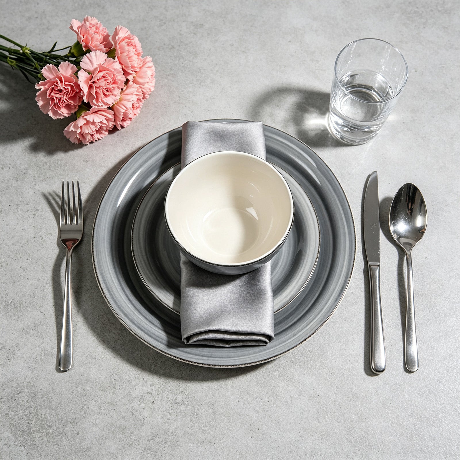 A styled tabletop place setting with a gray striped dinner plate, a white ceramic bowl, a gray napkin, silver fork and spoon, a knife, and a clear drinking glass on a light gray surface. Pink flowers appear in the upper left corner.