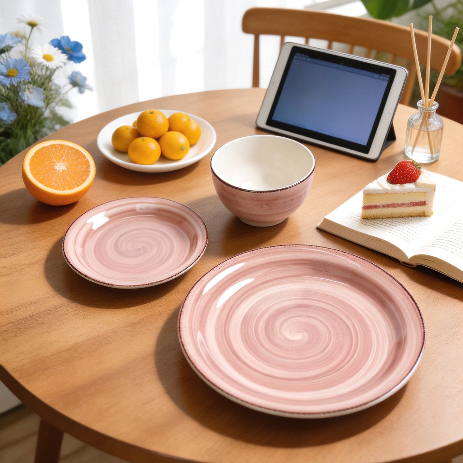 A styled dining table scene showing pink spiral ceramic plates and bowls, with an iPad, fruit, flowers, a book, and a slice of cake in a bright indoor setting.