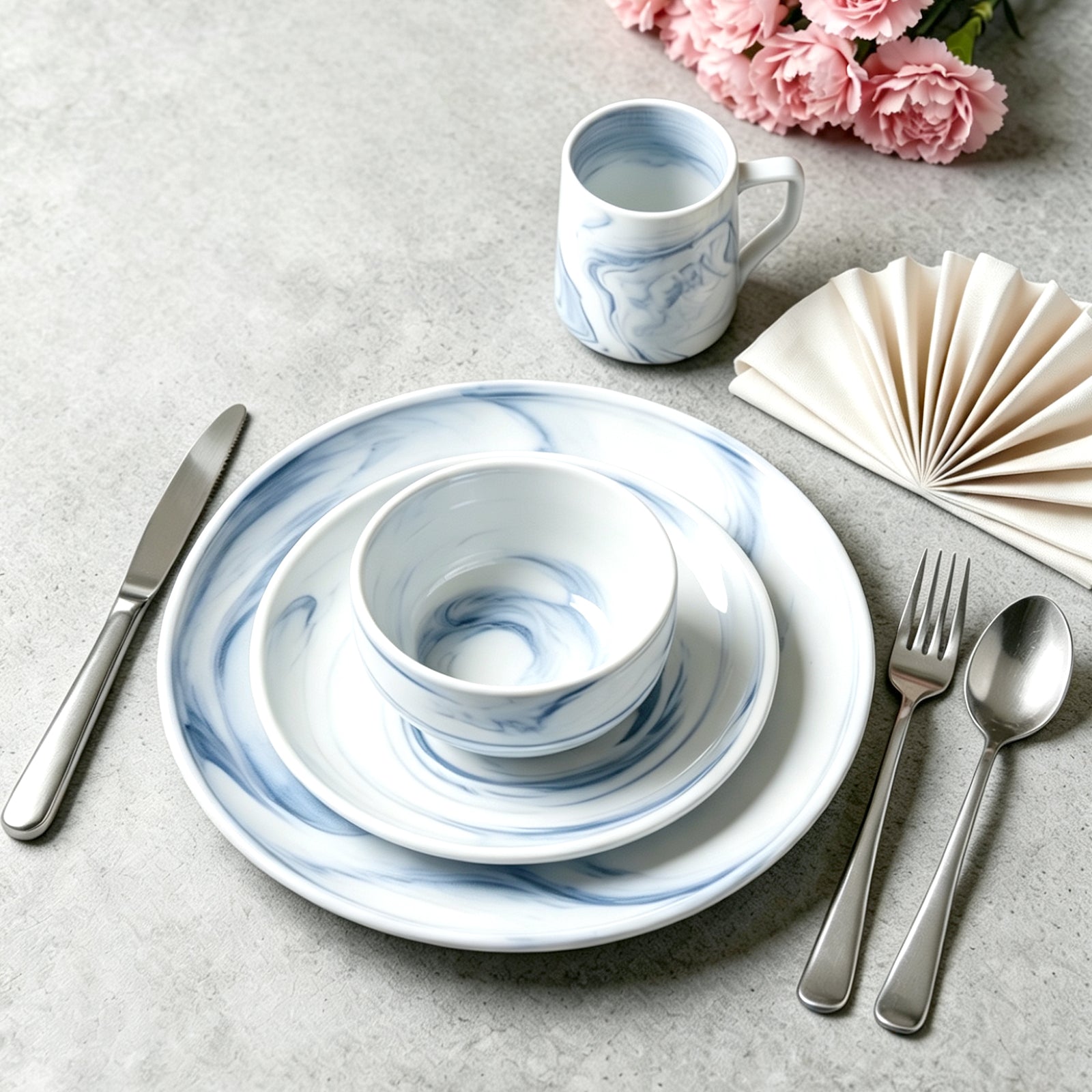 A place setting featuring a white ceramic dinnerware set with a blue marble swirl pattern, including a charger plate, bowl, and mug, styled with silver cutlery, a cream fan napkin, and pink flowers on a gray textured surface.