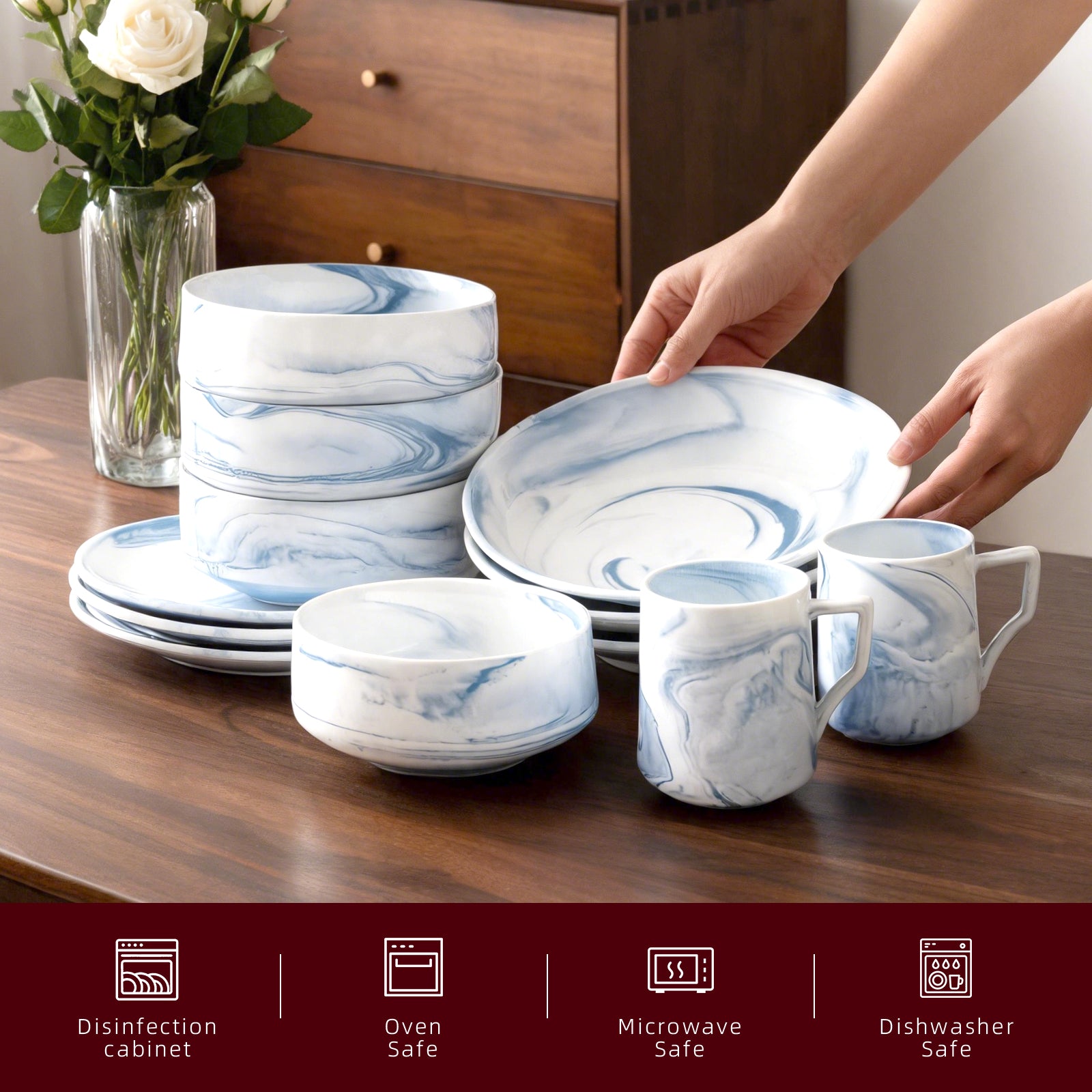 A set of white ceramic tableware with blue marble swirl patterns, including plates, bowls, and mugs, arranged on a dark wooden table. Two hands are visible holding a large plate, and a vase of flowers and a wooden cabinet are in the background.