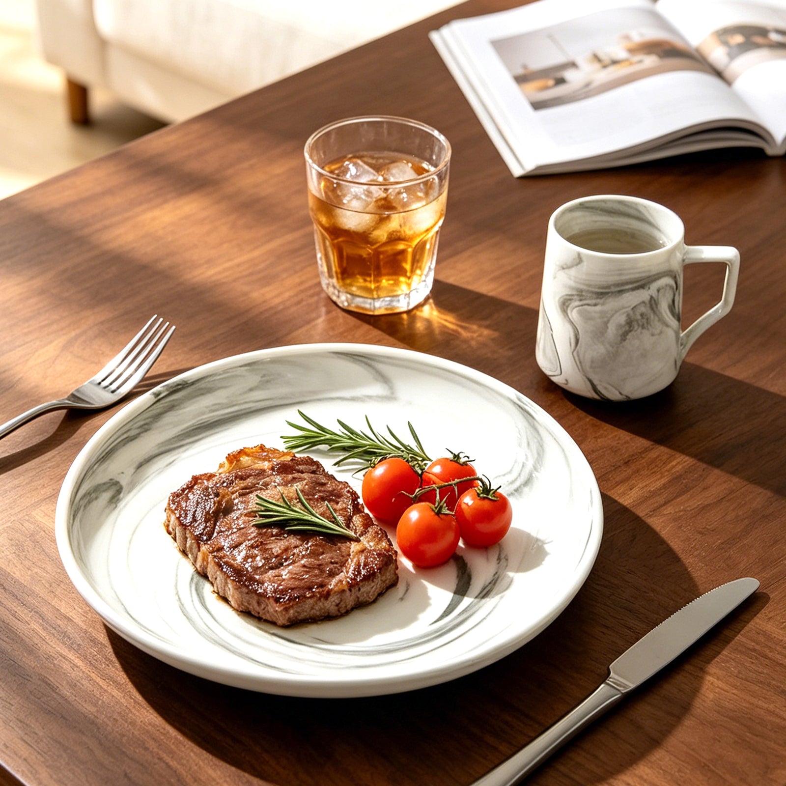 A white ceramic plate with a gray marble pattern is set on a wooden table, holding a cooked steak with rosemary and cherry tomatoes. A glass of iced amber drink, a gray-and-white marbled mug, a fork, and a knife are arranged around it, with an open magazine in the background.