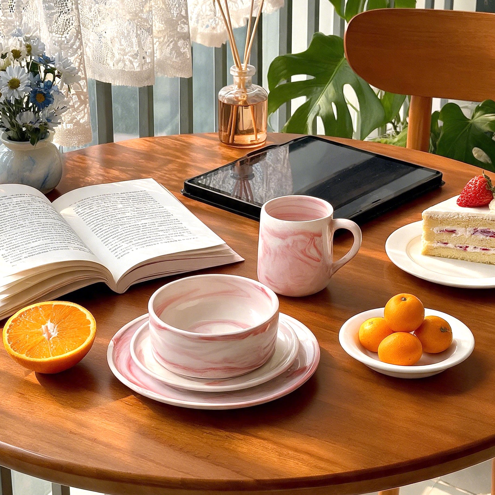 A styled dining table scene with a pink marbled ceramic bowl and matching saucer, a pink marbled mug, a tablet, an open book, fruit, and a slice of cake. No person is visible, but this is not a clean isolated studio product shot because it is set in a real environment with surrounding props and furniture, so subject_type is Lifestyle.