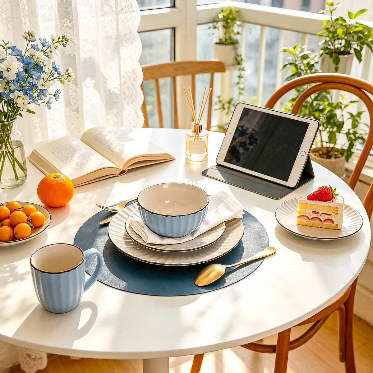 A beautifully styled round white dining table in a sunlit room, set with a blue mug, matching bowl and plates, a tablet in a black stand, a spoon, a book, oranges, a small cake with a strawberry, and flowers in the background.