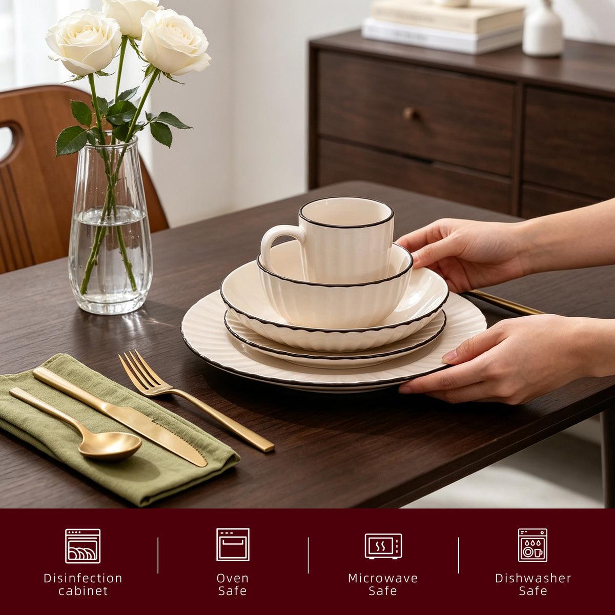 A set of beige ceramic dinnerware with black rims, including a plate, bowl, and mug, being placed on a dark wood table by hands. The table is styled with a green napkin, gold-toned cutlery, a glass vase of white roses, and a matching gold utensil.