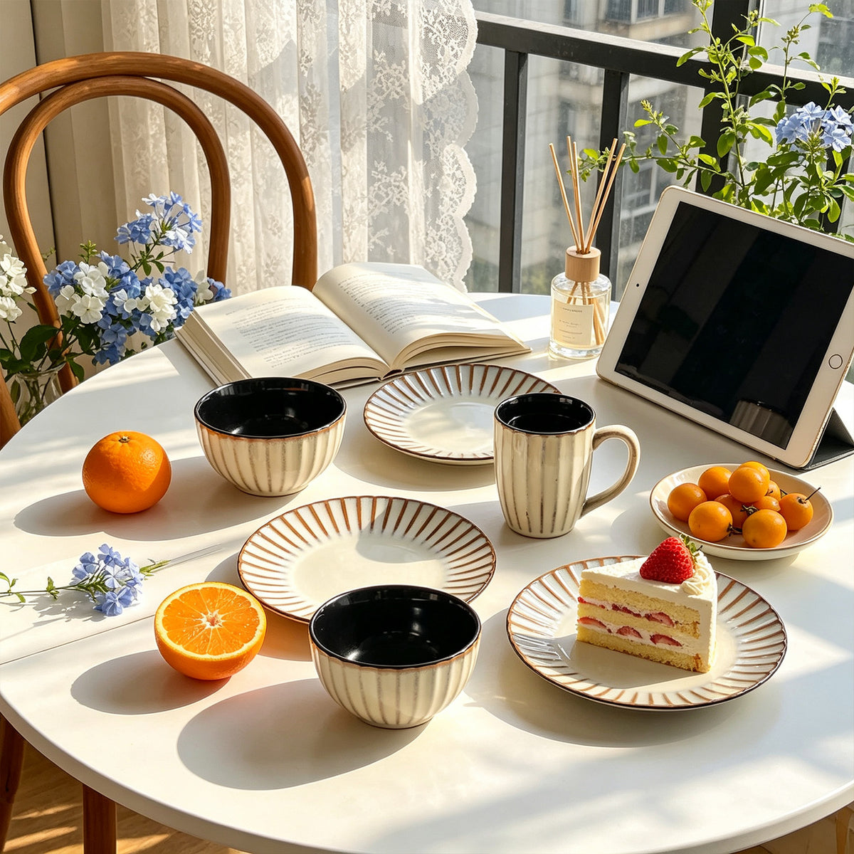A styled tabletop scene with cream and black ceramic tableware, including bowls, plates, and mugs, arranged on a round white table. An open book, fruit, a slice of cake, a tablet, flowers, and a chair are visible in a bright indoor setting with sunlight and window views.