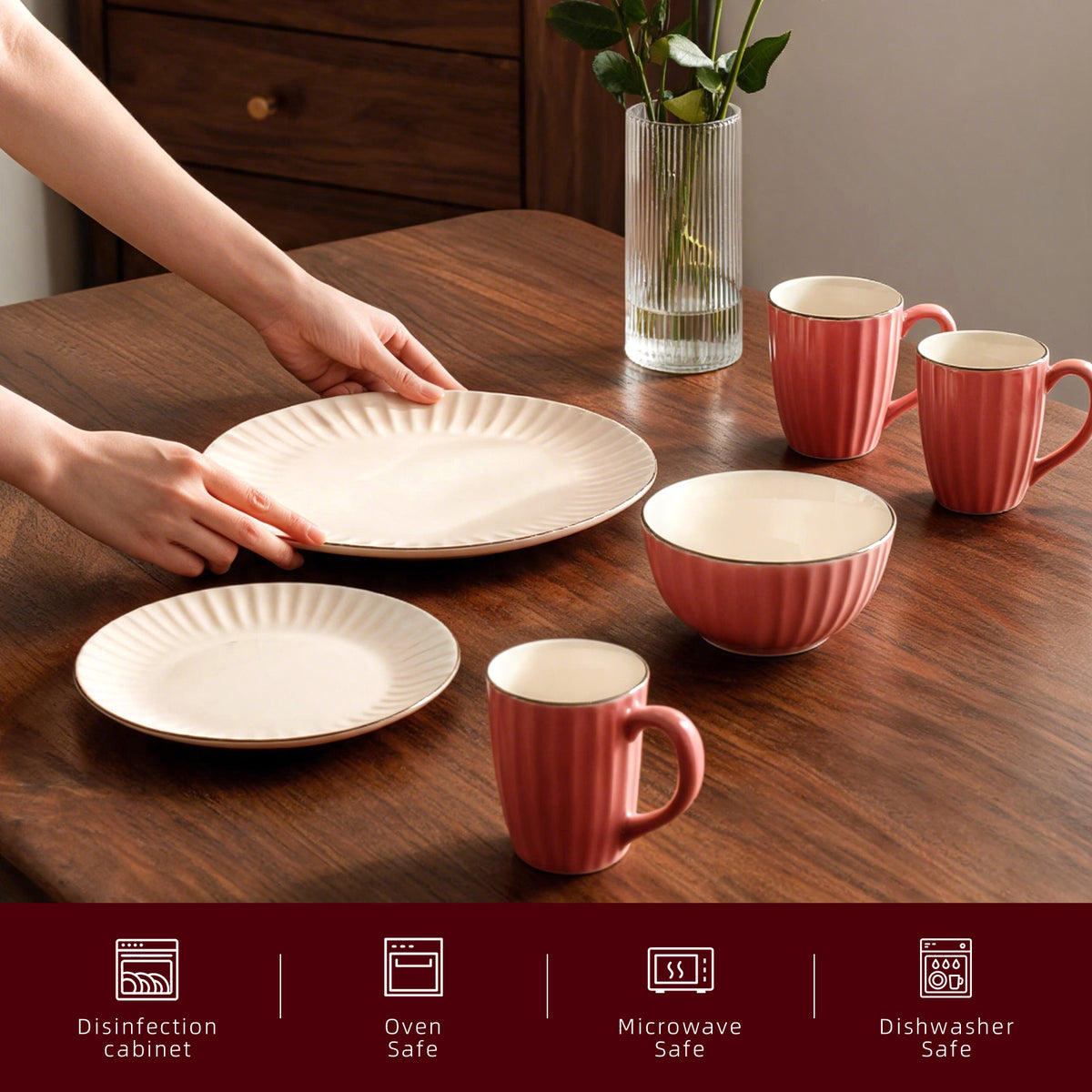A set of ceramic dinnerware including plates and mugs in a warm pinkish-red ribbed finish with cream interiors, arranged on a wooden dining table. A person's hands are visible holding one plate, and a glass vase with green leaves sits in the background.
