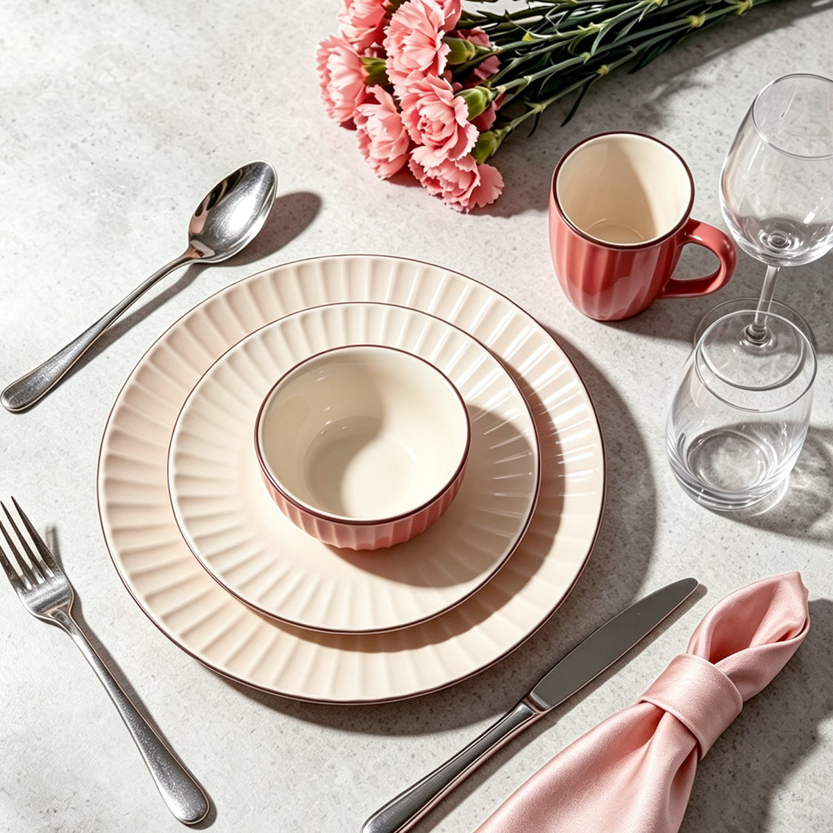 A styled table setting featuring a pink and cream ceramic dinnerware set, including a charger plate, a bowl, and a mug, arranged on a light gray textured surface with silver cutlery, a pink napkin, and pink flowers.