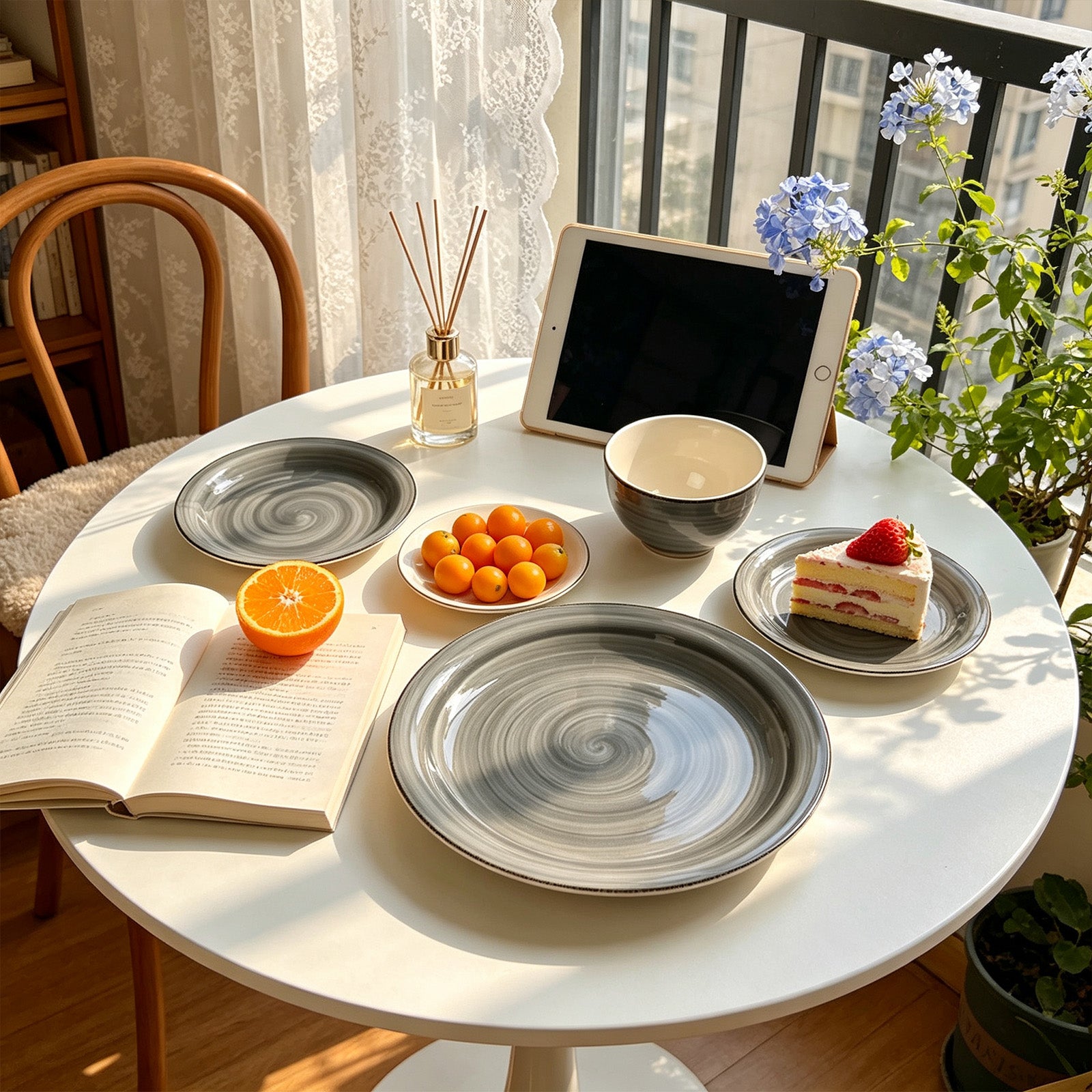 A styled dining table scene with gray and white ceramic plates, a bowl, a tablet on a stand, an open book, an orange half, small orange fruits, and a slice of cake, set in a bright indoor environment with plants and a chair.