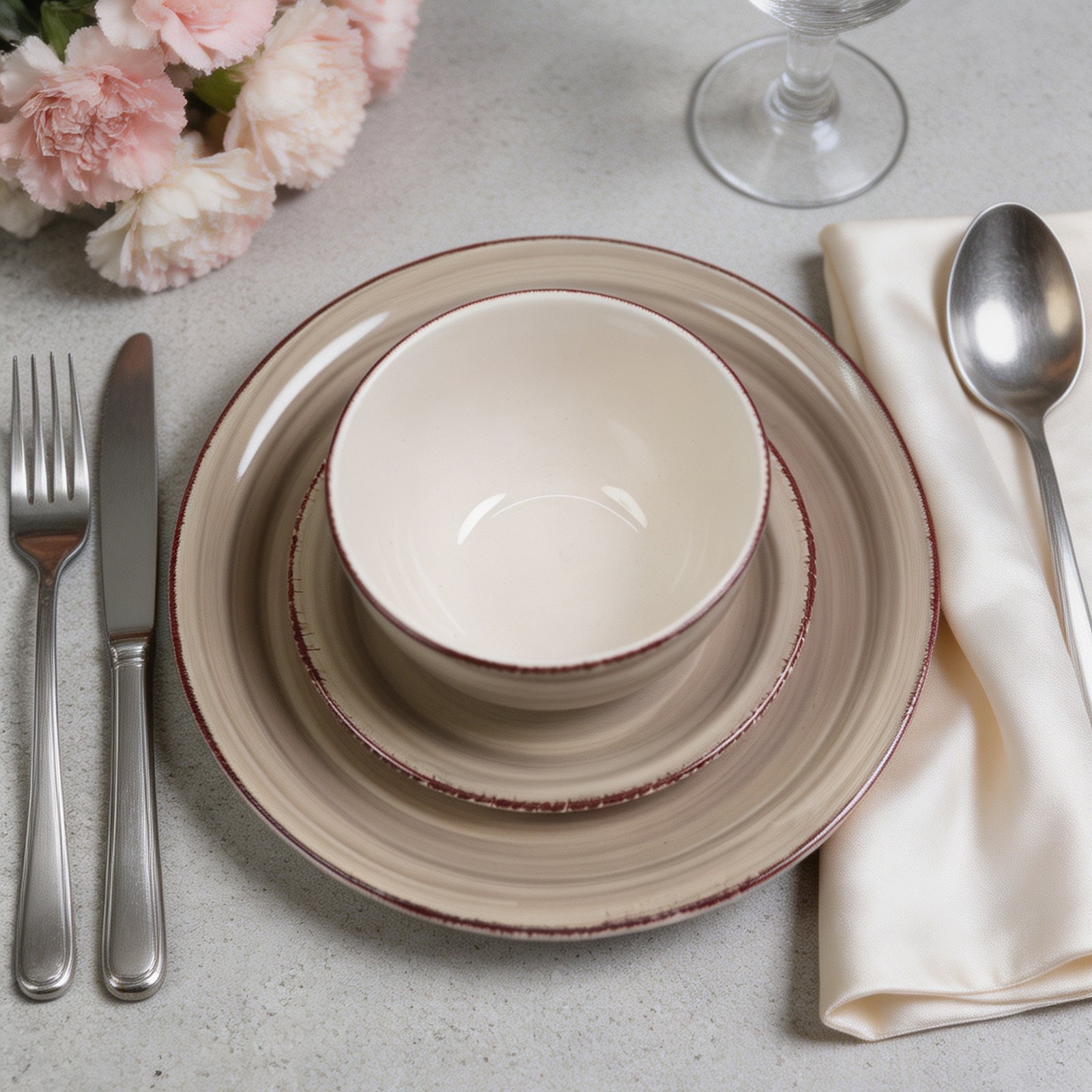 A place setting featuring a ceramic dinnerware stack with a beige bowl, saucer, and charger plate with a rustic reddish-brown rim, styled with silver cutlery, a cream napkin, and pink flowers on a light gray surface.