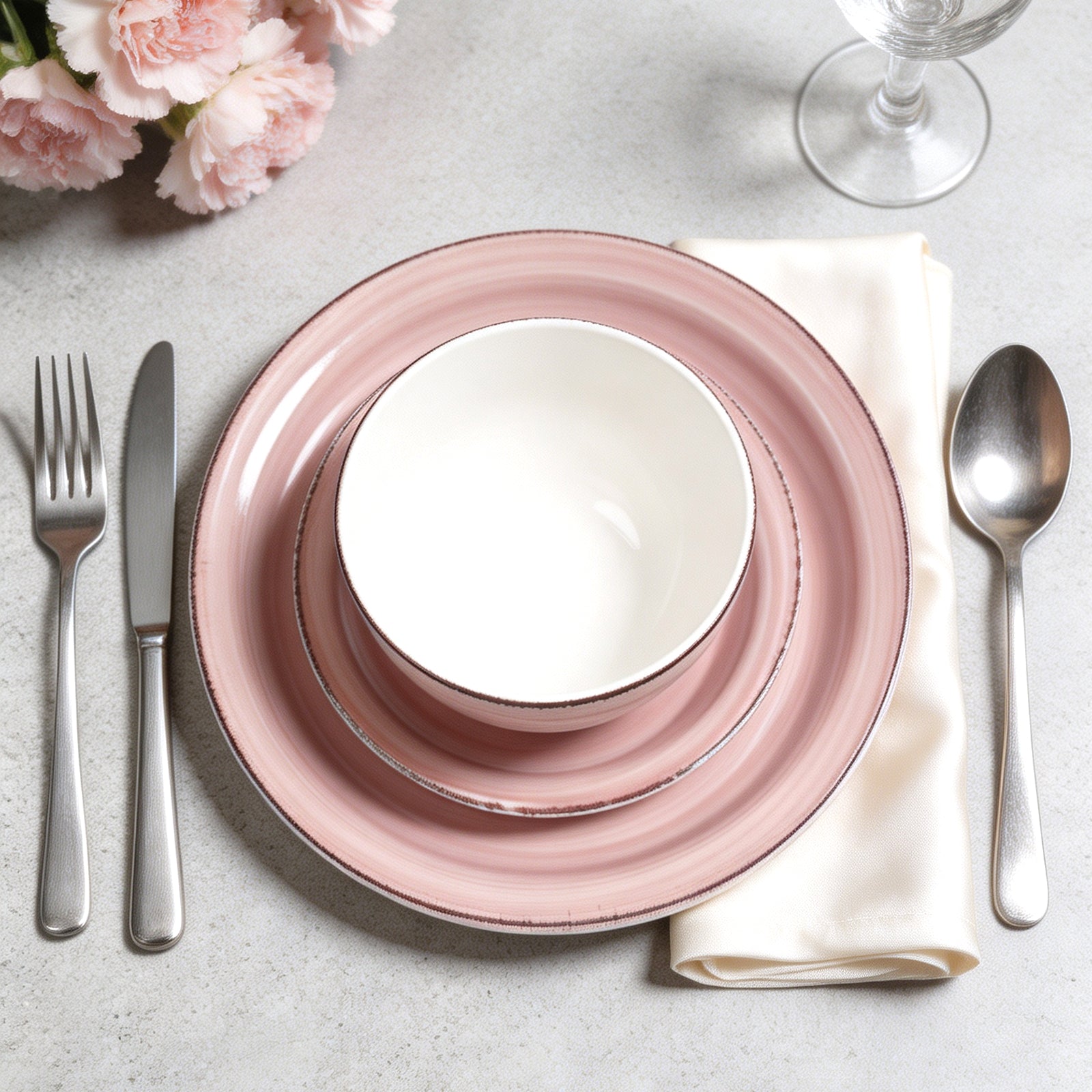 A place setting featuring a pink ceramic dinnerware set with a white bowl, accompanied by silver cutlery and a cream napkin on a light gray textured surface with pink flowers in the background.