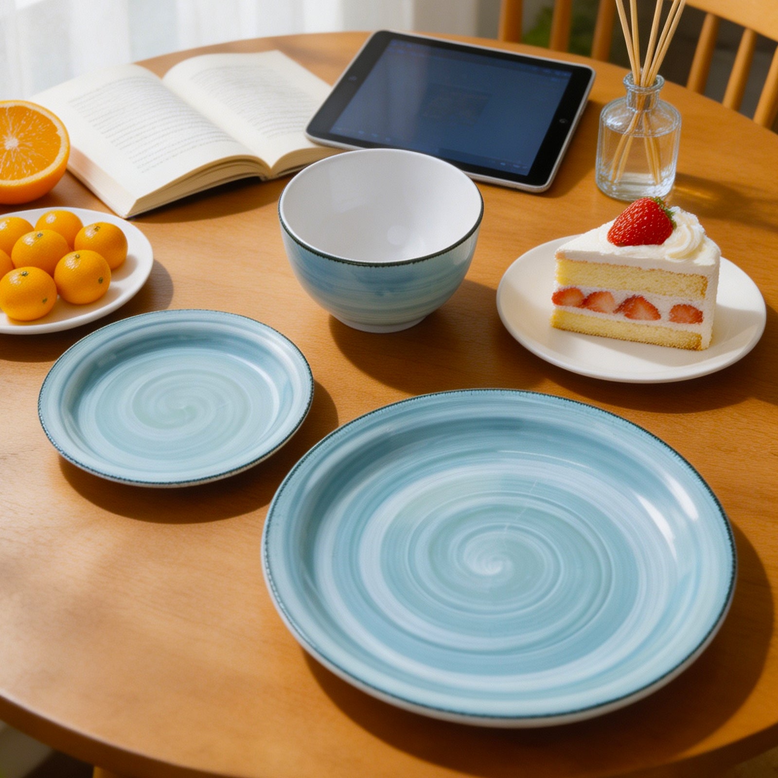 A set of light blue and white ceramic tableware, including a large plate, a smaller plate, and a bowl, styled on a wooden dining table with an open book, oranges, a tablet, and a slice of cake.