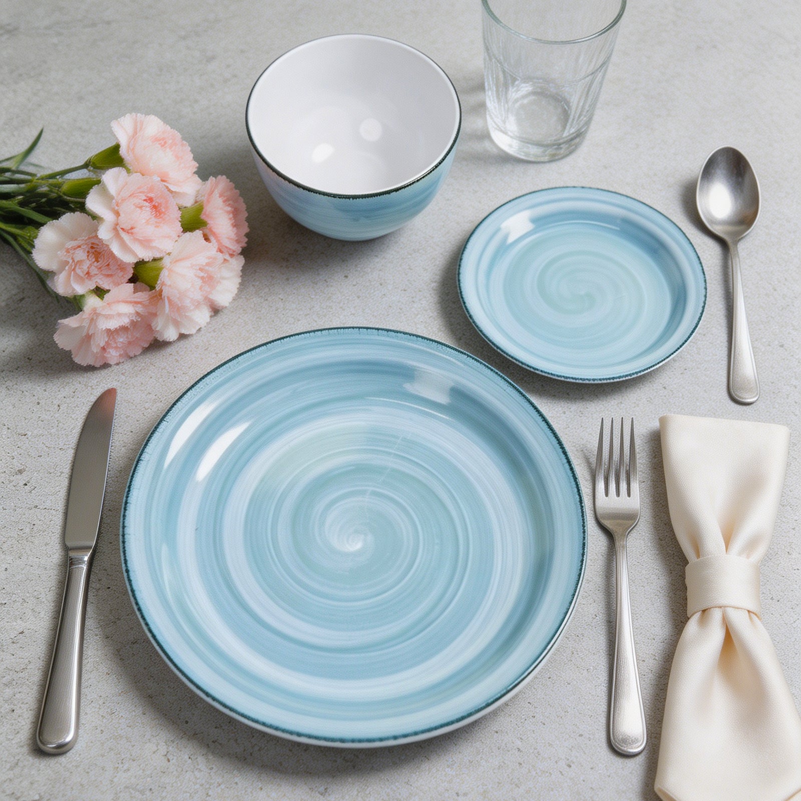 A light blue and white spiral-patterned dinnerware set arranged on a light gray surface, including a large plate, a smaller plate, a bowl, a glass, silver cutlery, a cream napkin, and pink flowers.