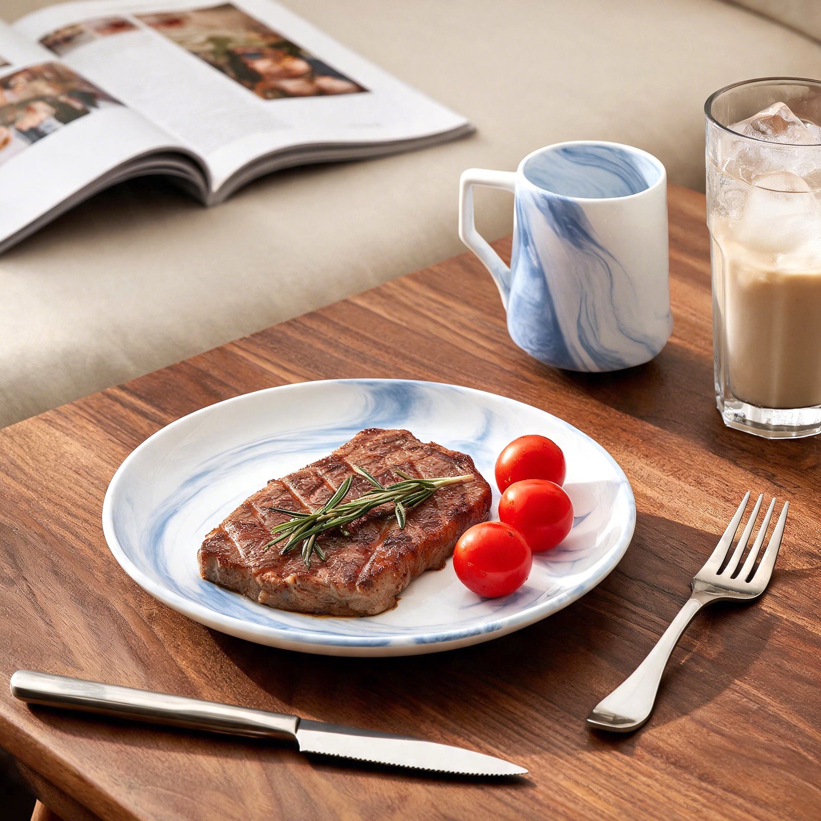 A styled dining table scene featuring a white ceramic plate with a blue marble pattern, holding a grilled steak with a rosemary sprig and two cherry tomatoes. A matching blue-and-white marbled mug, a glass of iced coffee, a fork, and a knife are arranged on a wooden table, with an open magazine in the background.