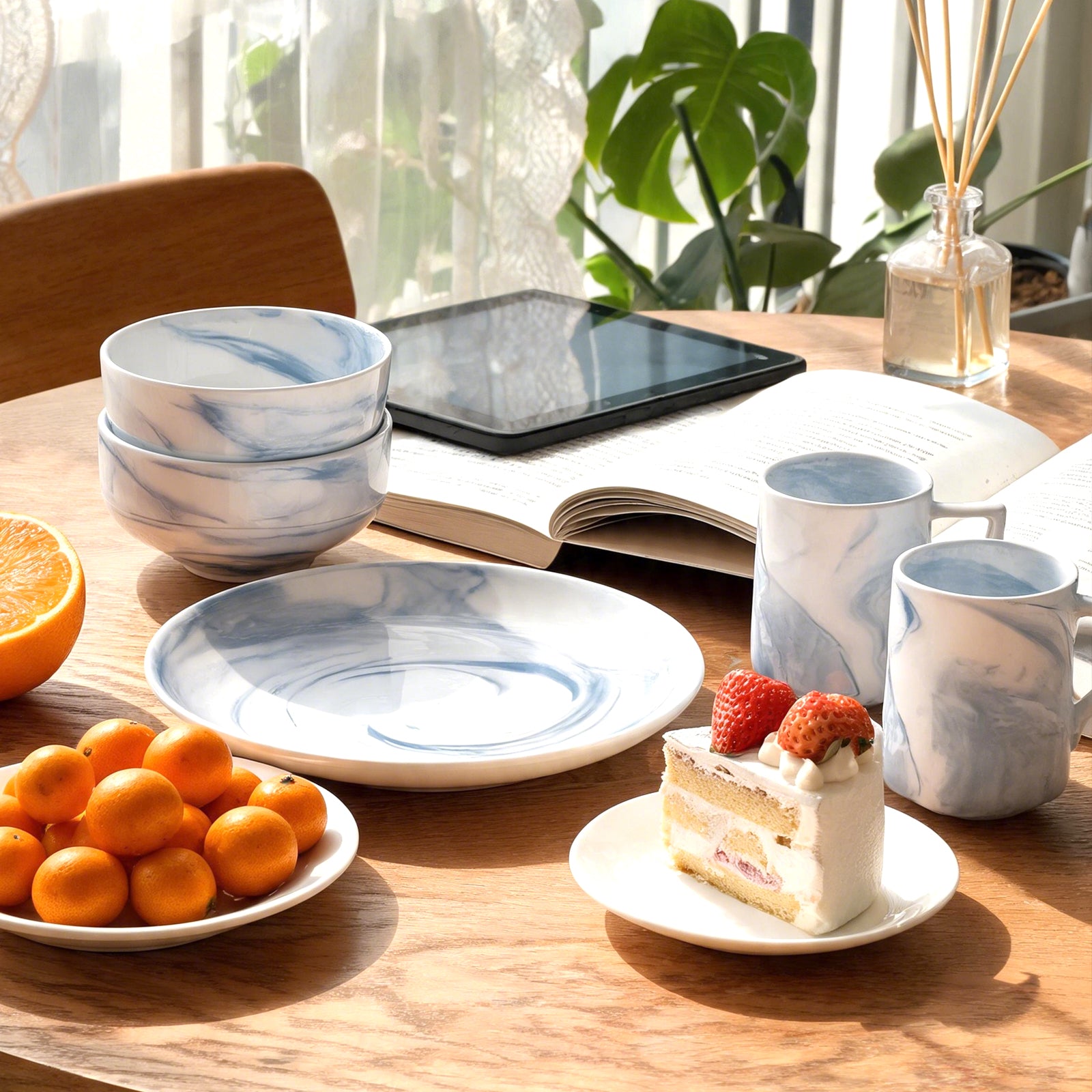 A styled tabletop scene with a set of white ceramic tableware featuring a blue marble pattern, including bowls, plates, and mugs, arranged on a wooden table with books, a tablet, fruit, and a slice of cake.