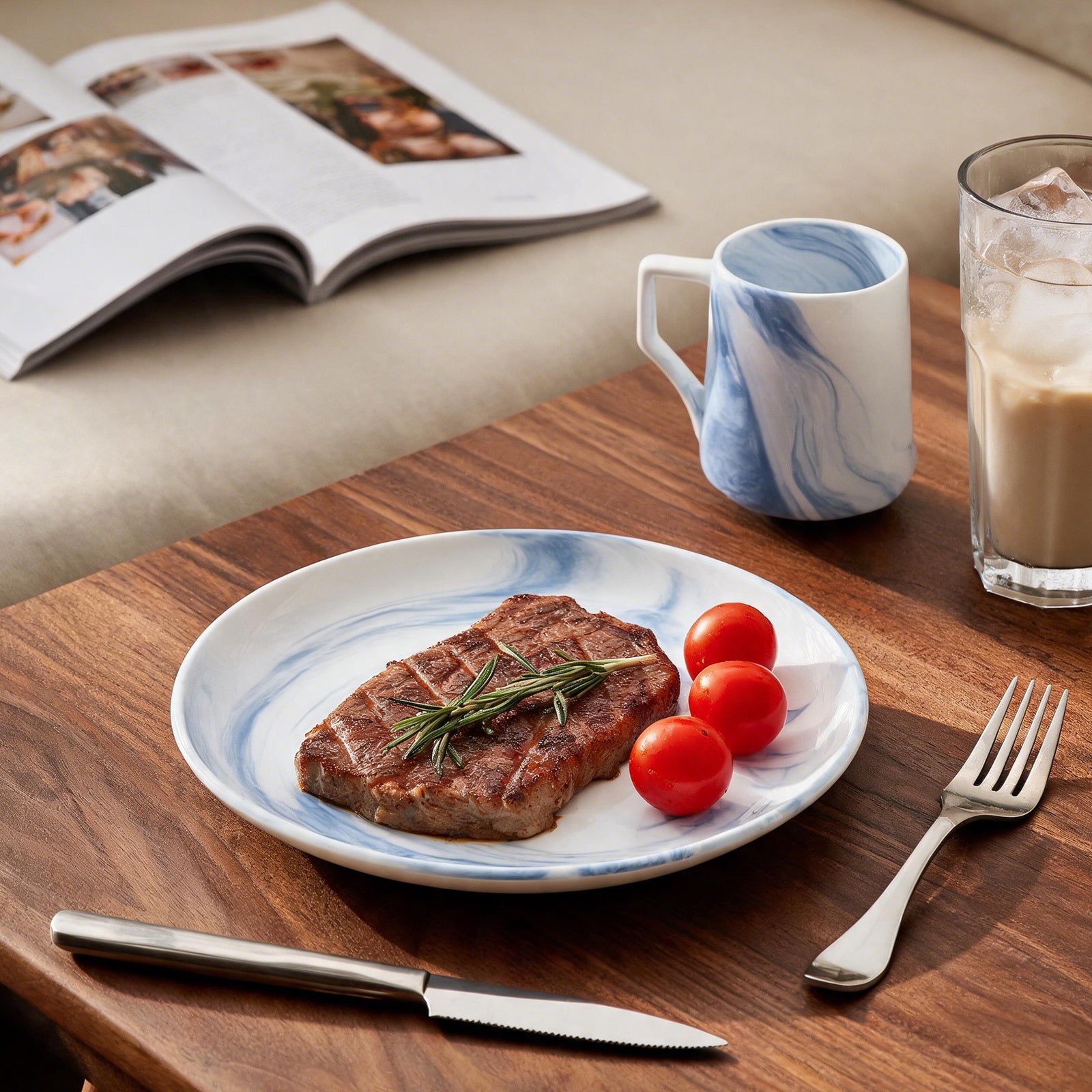 A round white ceramic plate with a blue marble pattern, styled with a grilled steak, cherry tomatoes, a knife, and a fork on a wooden table. In the background, there is an open magazine and a blue-and-white marbled mug.