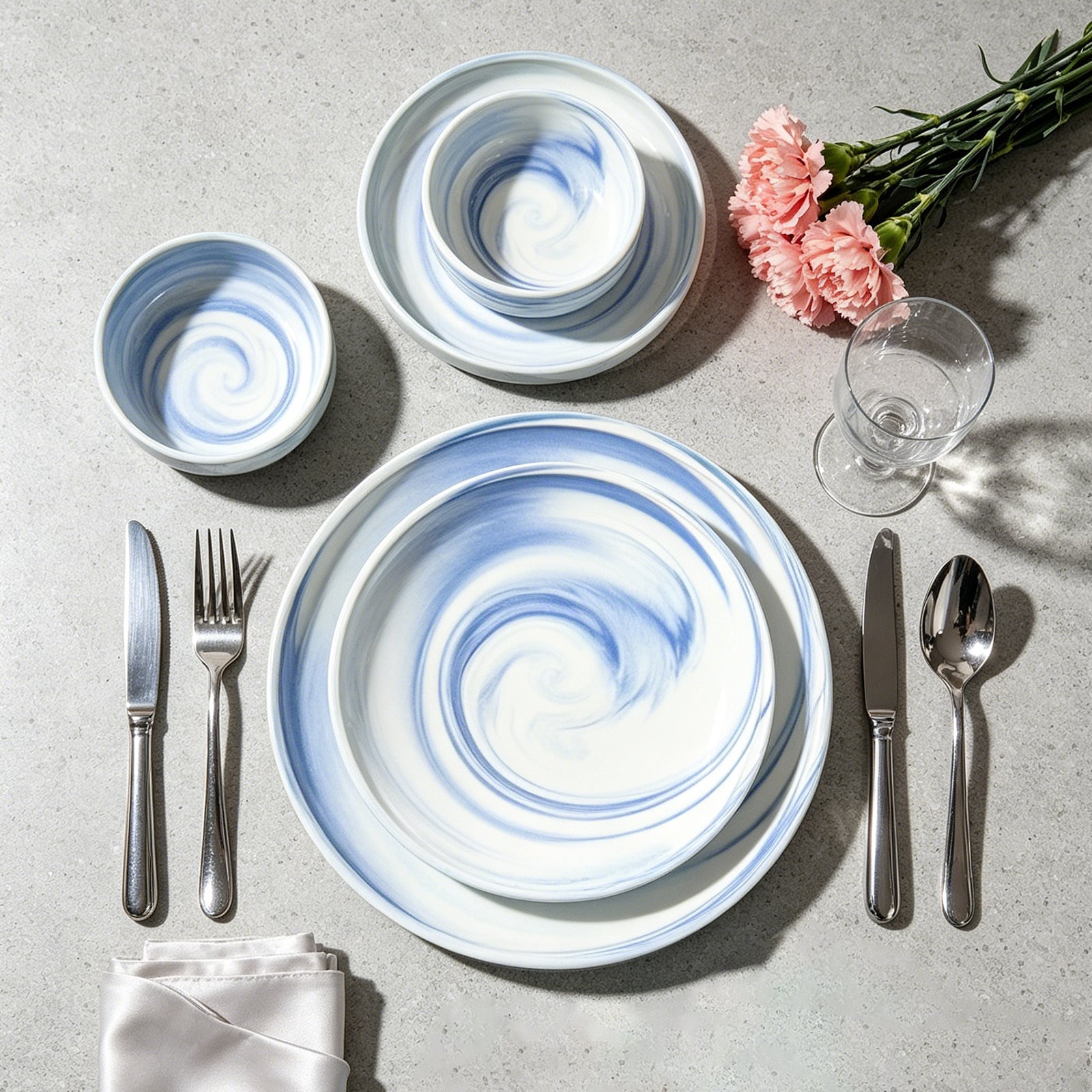 A styled flat-lay of a table setting featuring a matching dinnerware set with a white and blue marbled swirl pattern. The set includes a large plate, a charger plate, two bowls, and silver cutlery on a light gray textured surface, accented with pink carnation flowers and a glass.