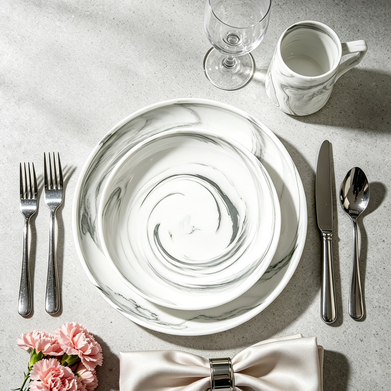 A styled tabletop setting featuring a white ceramic plate with a gray marble swirl pattern, accompanied by silver cutlery, a matching white mug, a wine glass, and pink flowers on a light gray surface.