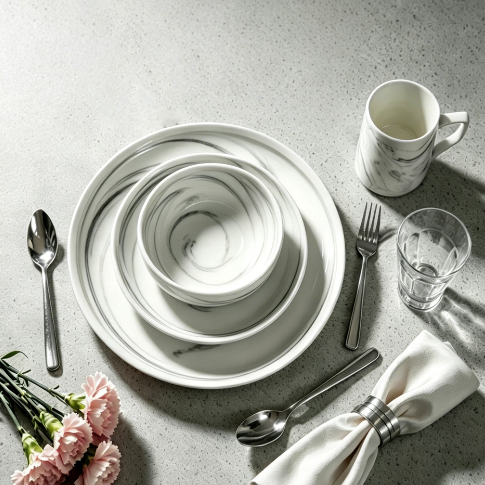 A styled tabletop setting with a white ceramic dinnerware set featuring a gray marble swirl pattern, including plates, bowls, and a mug, arranged on a light gray textured surface with silver cutlery, a glass, a folded cream napkin, and pink flowers.
