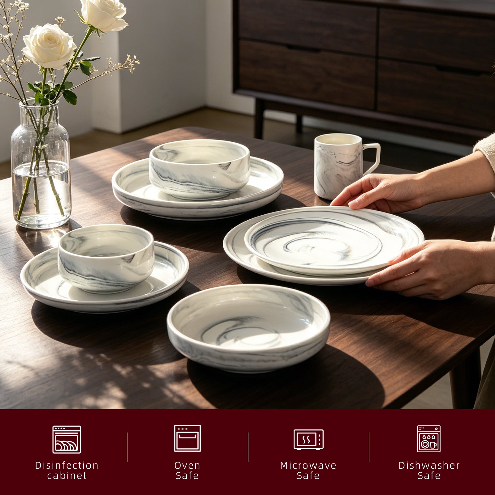 A set of white ceramic tableware with a gray marble pattern, including plates, bowls, and a mug, arranged on a dark wooden dining table. A person's hands are visible placing a plate, and a glass vase with white roses sits on the left.