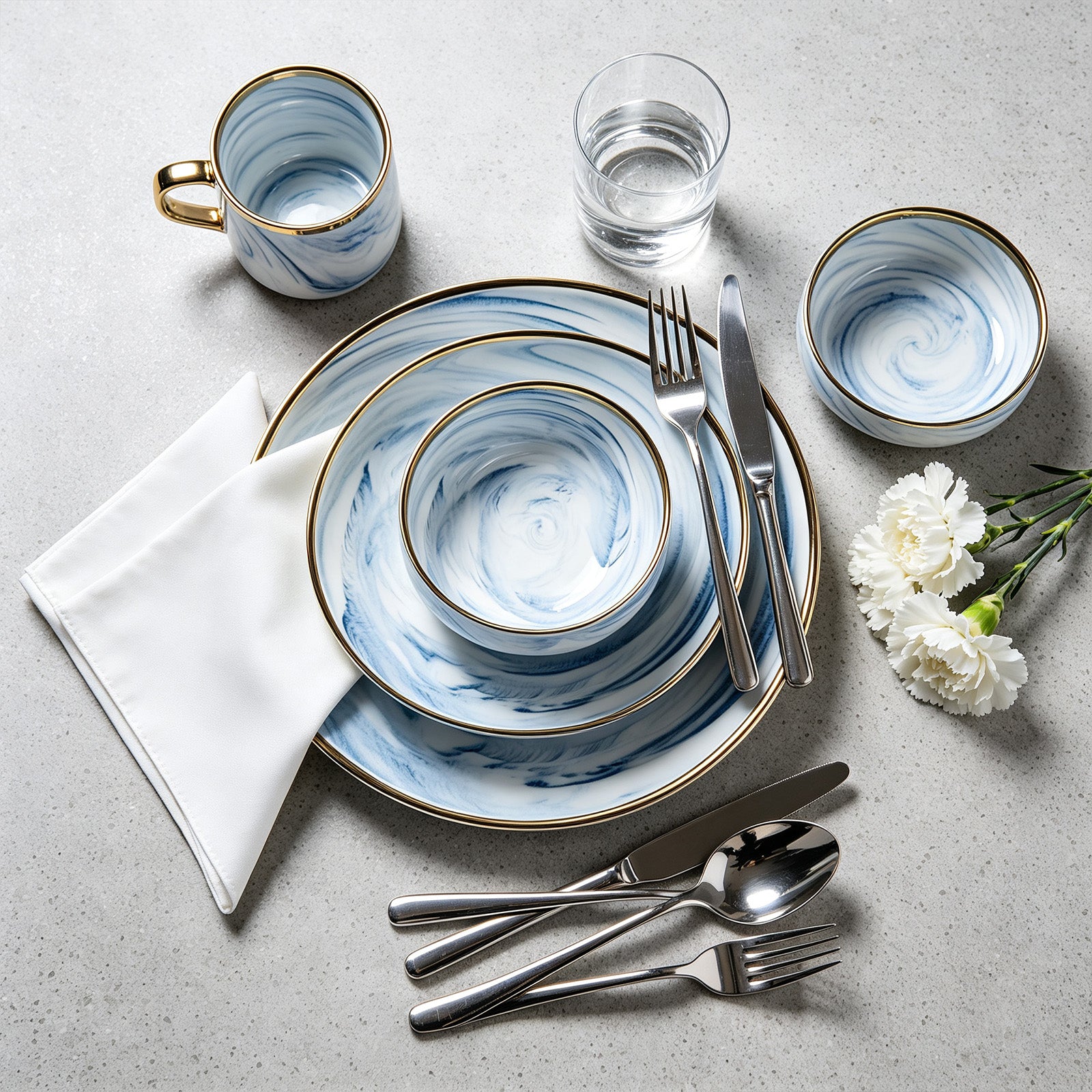 A place setting featuring a coordinated blue and white marbled dinnerware set with gold trim, arranged on a light gray textured surface with silver cutlery, a glass of water, and white flowers.