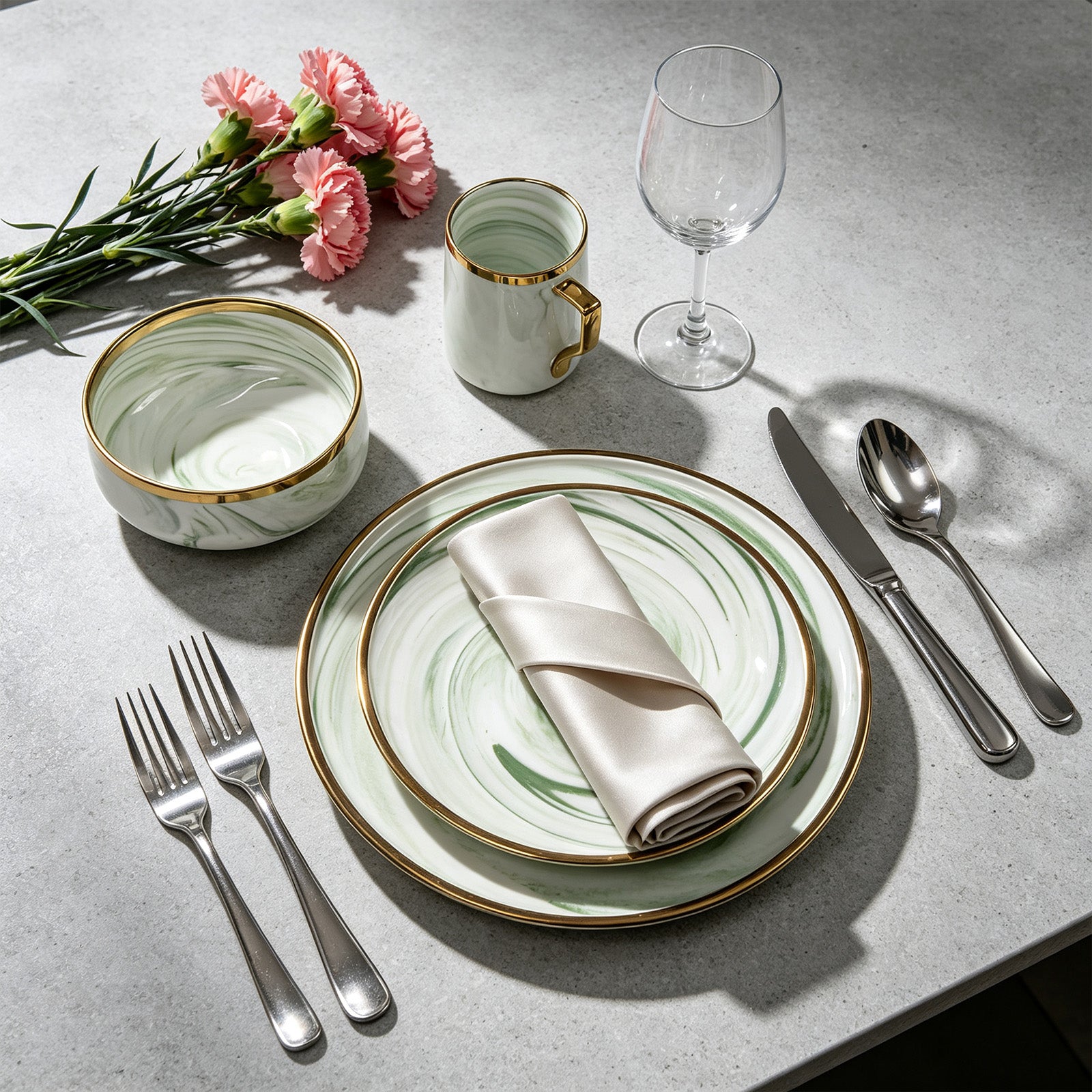 A styled place setting on a light gray stone surface featuring a green and white marbled dinner plate set with gold trim, a matching bowl, a folded cream napkin, silver cutlery, a clear wine glass, and a small mint-green mug with gold trim, accented by pink carnation flowers.