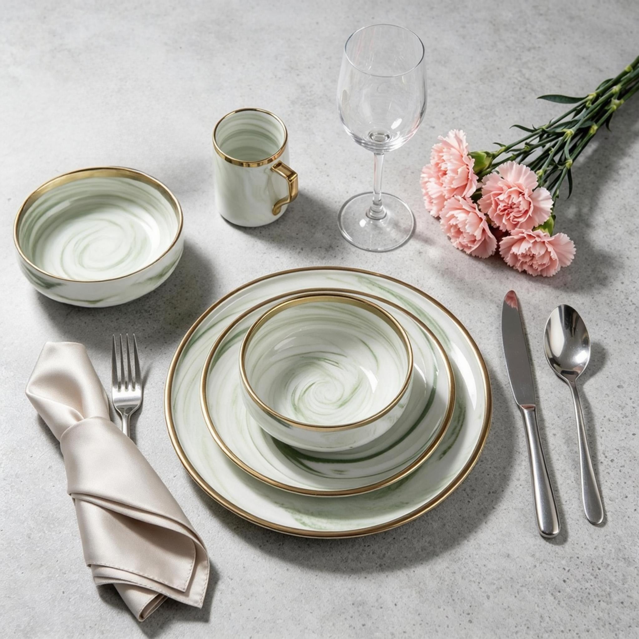 A place setting featuring a stack of white ceramic plates and bowls with elegant gold rims and a subtle green swirl pattern, accompanied by a matching cup, silver cutlery, a folded cream satin napkin, and pink carnation flowers on a textured gray surface.