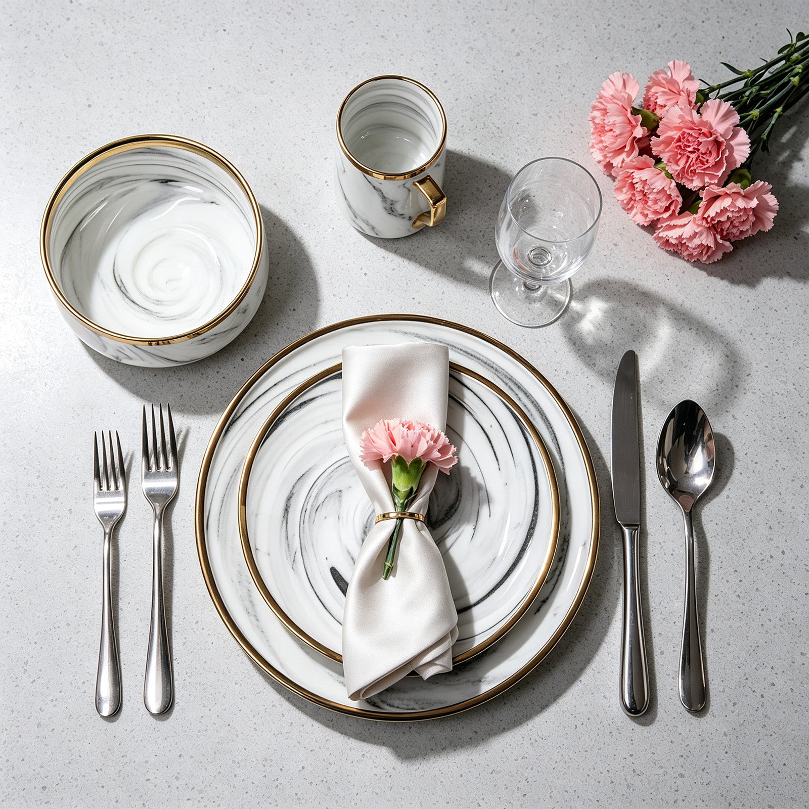 A styled flat-lay of a place setting on a light gray stone surface, featuring a white and gray marbled dinner plate with a gold rim, a matching bowl, a small gold-handled cup, a clear wine glass, silver-toned cutlery, a folded white napkin with a pink carnation, and a bouquet of pink carnations.