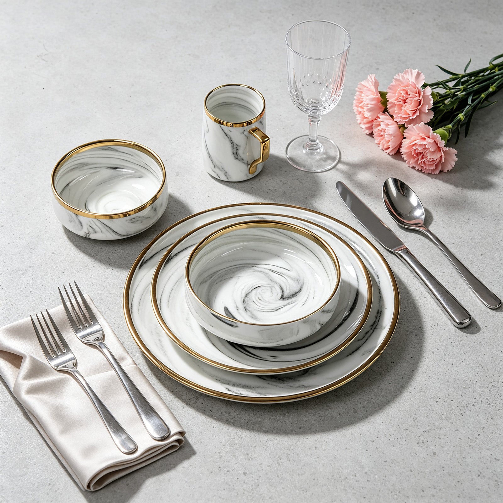 A place setting featuring a marble-patterned dinnerware set with gold trim, including plates, bowls, a mug, a glass, silver cutlery, a folded cream napkin, and pink carnation flowers on a light gray textured surface.