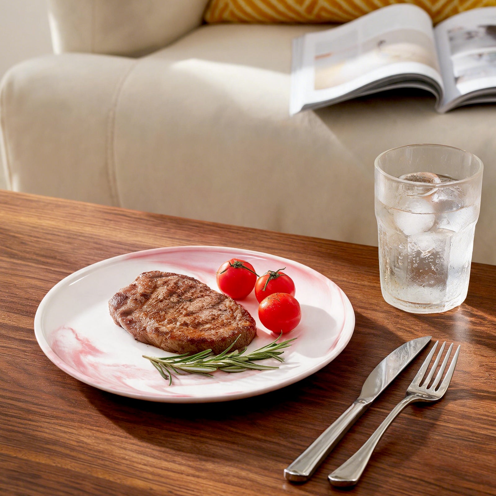 A white ceramic plate with a pink floral border, styled with a grilled steak, cherry tomatoes, and rosemary on a wooden table. A glass of water, a knife, and a fork are placed beside it, with a beige sofa and an open magazine in the background.