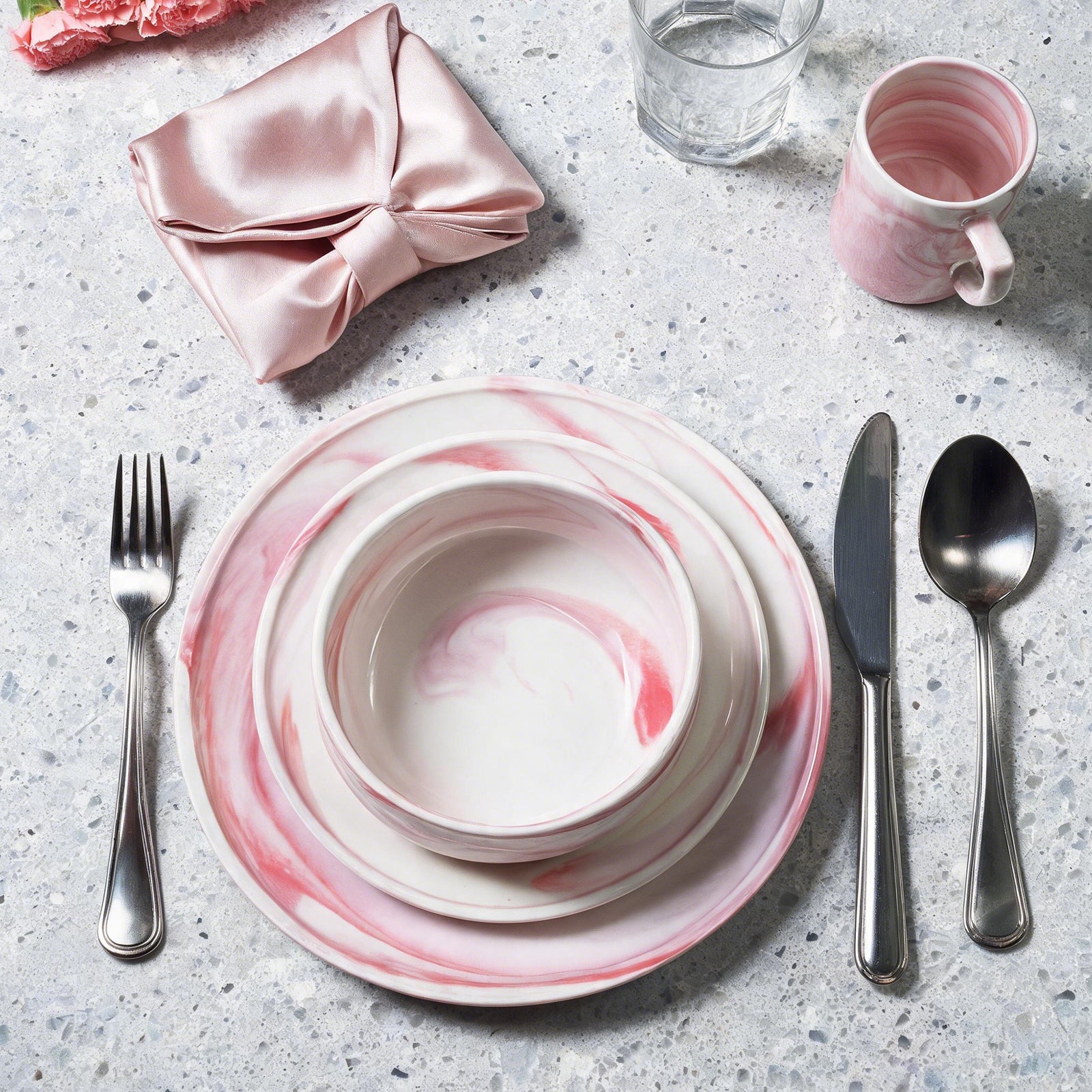 A place setting featuring a stack of white ceramic plates with a pink marble swirl pattern, accompanied by silver-toned flatware and a matching pink ceramic mug on a speckled gray countertop.