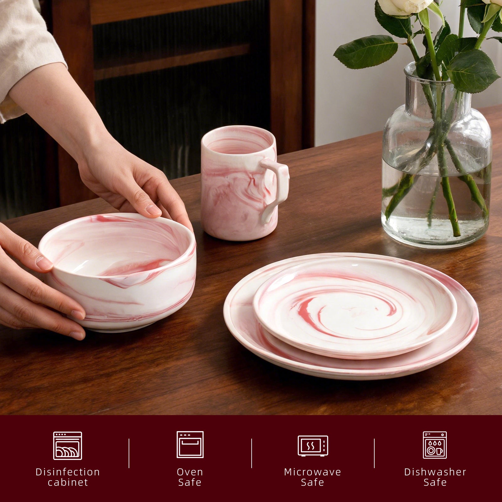 A set of pink and white marbled ceramic tableware, including a bowl, a mug, and stacked plates, arranged on a dark wooden table. A person's hands are visible holding the bowl, and a glass vase with flowers sits in the background.