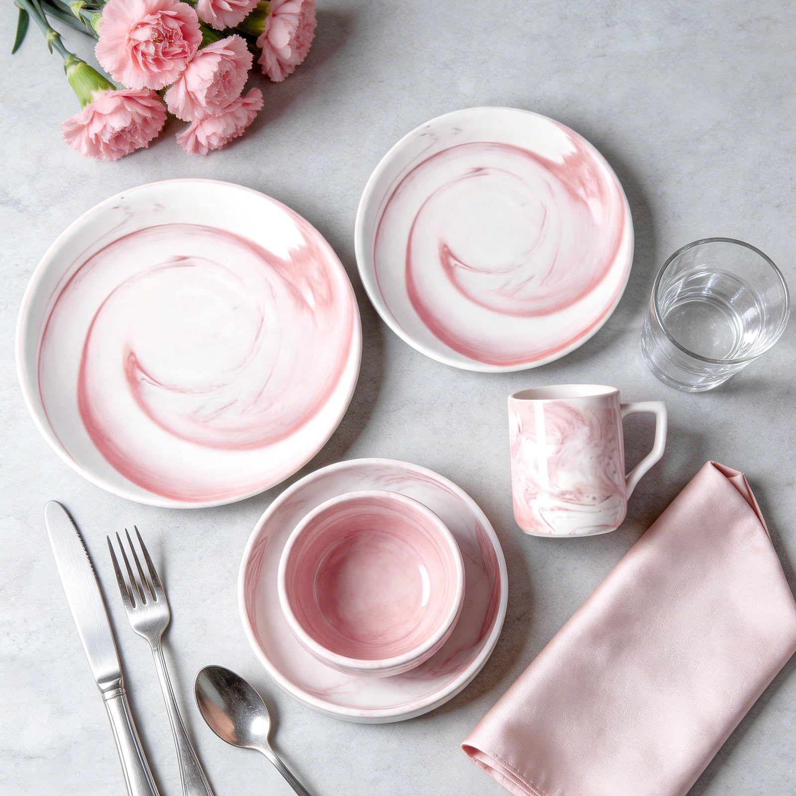 Pink and white marble-patterned dinnerware set arranged on a light gray surface with silver cutlery, a glass of water, pink flowers, and a folded pink napkin.