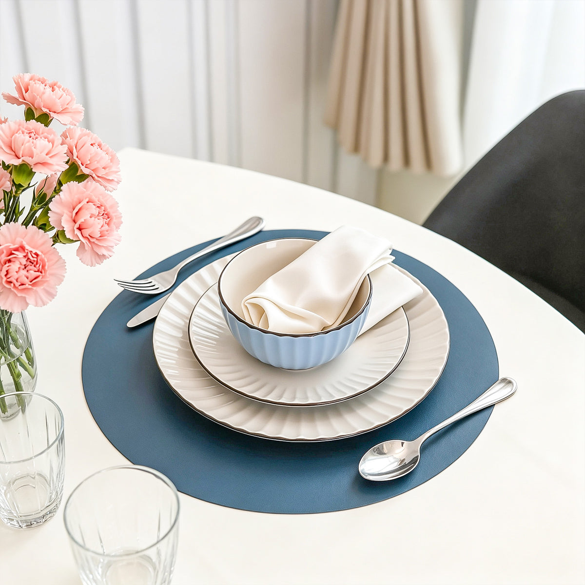 A styled dining table setting featuring a blue circular placemat, white plates, a light blue ribbed bowl with a cream napkin, silver cutlery, and a vase of pink carnations in the background.