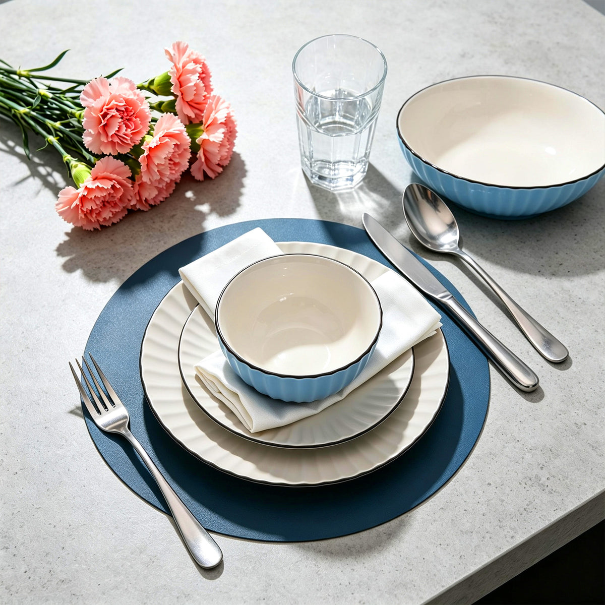 A styled table setting with a light blue and white ceramic bowl, matching plates, silver cutlery, a glass, and pink carnation flowers on a light gray surface.