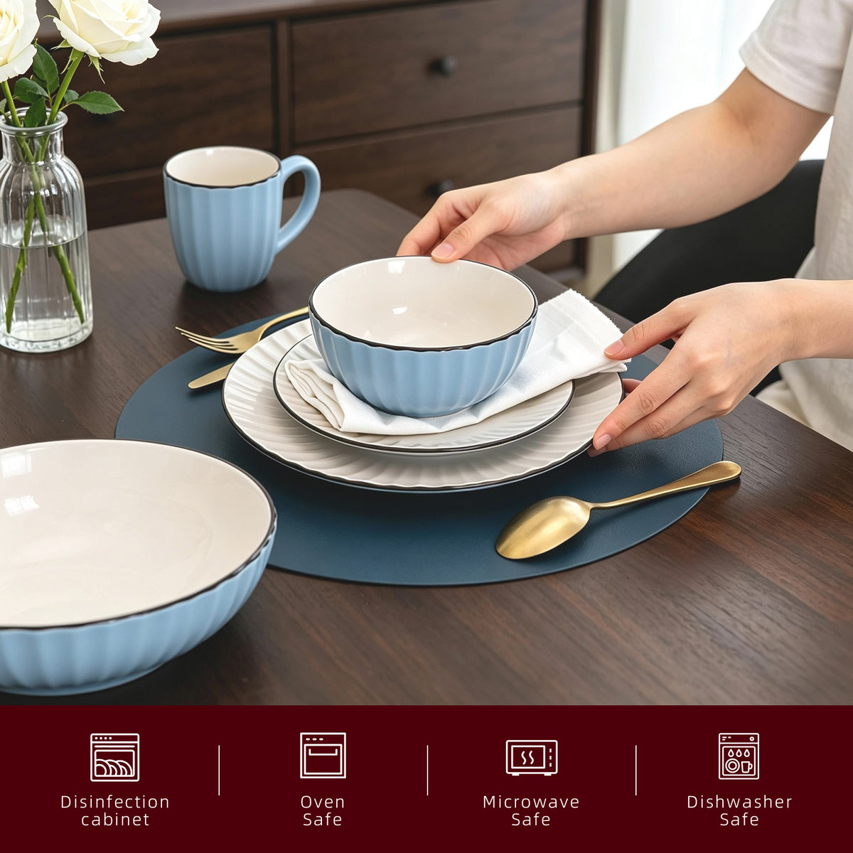 A light blue and white ceramic dinnerware set on a dark wood table, with a person's hands adjusting a bowl on a stack of plates. The set includes bowls, plates, a mug, and gold-toned cutlery, arranged with placemats and a vase of white roses.