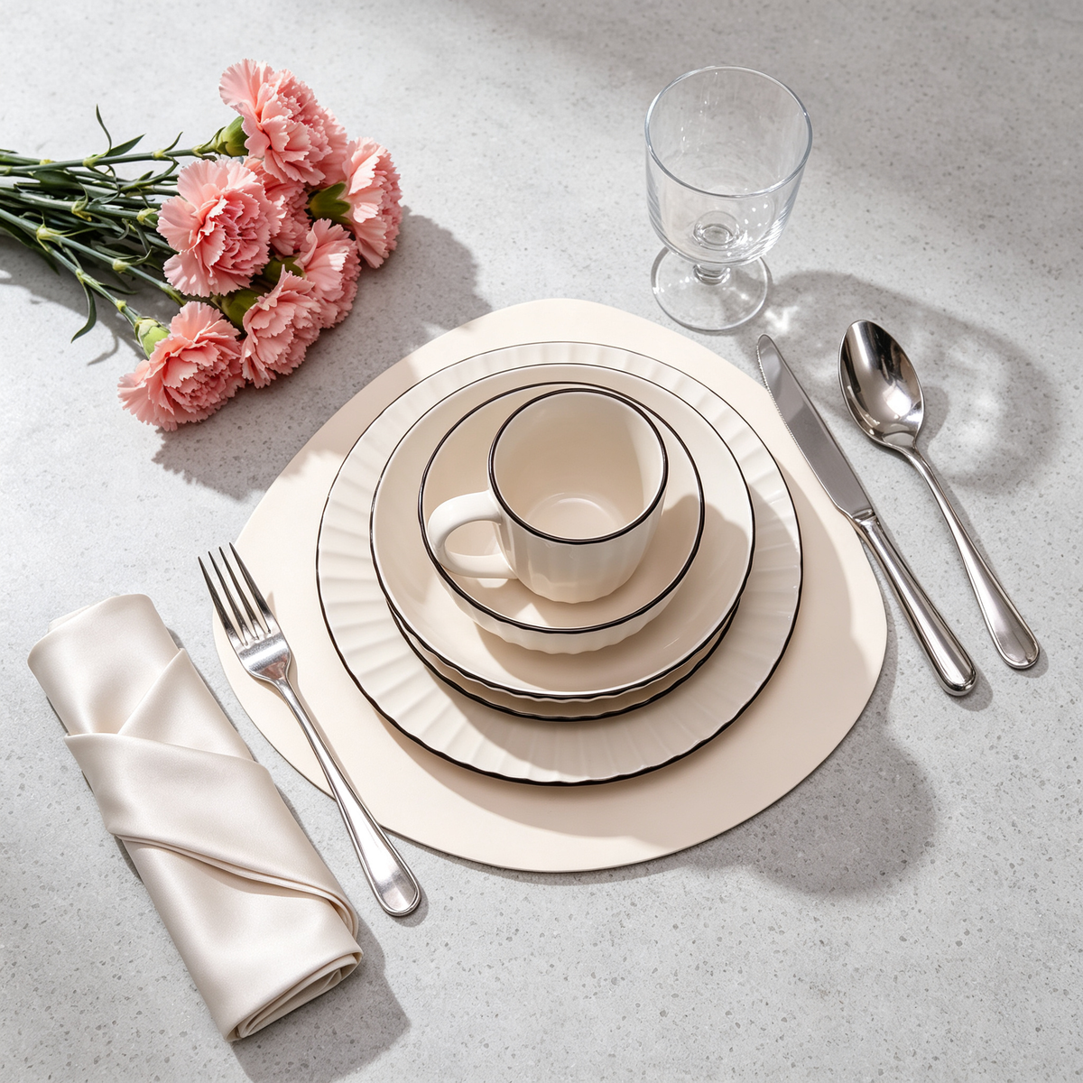 A styled table setting with a cream-colored ceramic dinnerware set, including a cup, plates, and a folded napkin, arranged on a light gray textured surface alongside silver cutlery, a clear drinking glass, and pink carnation flowers.