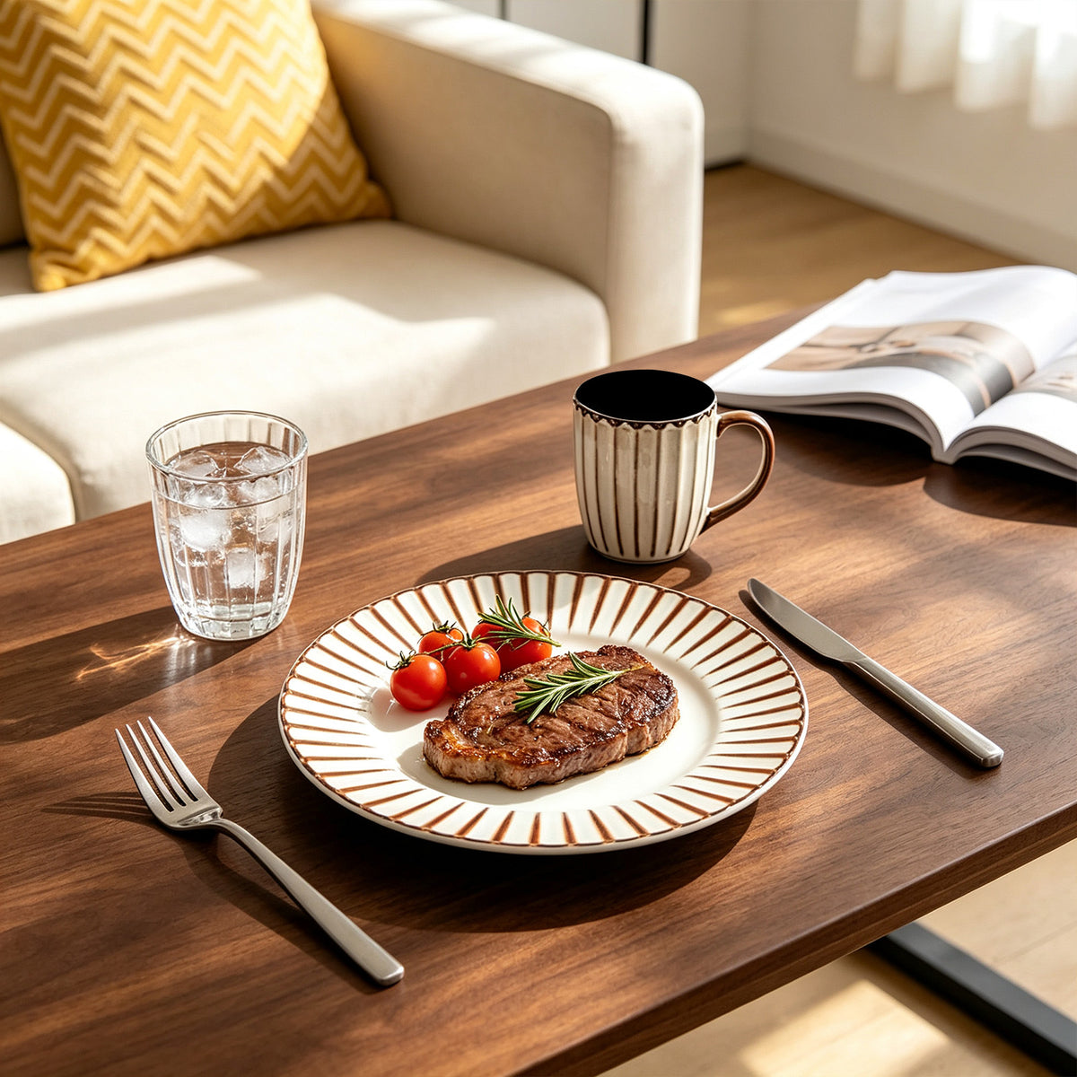 A styled dining table scene in a bright living space, featuring a round white plate with brown radial stripes holding a cooked steak with cherry tomatoes and a rosemary sprig, alongside a striped mug, glass of water, knife, fork, and an open magazine. A cream sofa with a yellow patterned pillow is visible in the background.