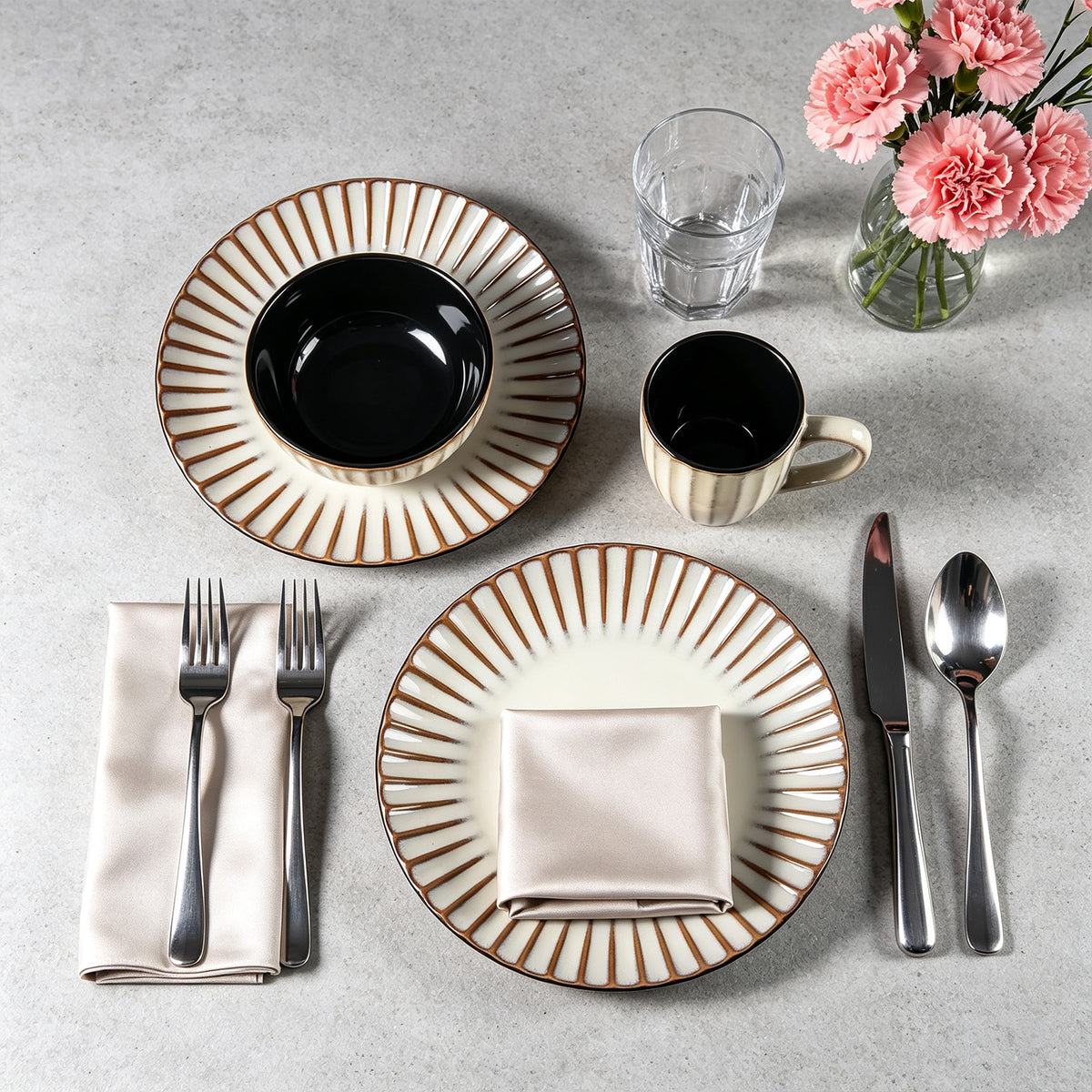 A styled flat-lay of a table setting on a light gray surface, featuring cream and brown striped ceramic plates, bowls, a mug, silver cutlery, a clear drinking glass, and pink flowers in a glass vase.