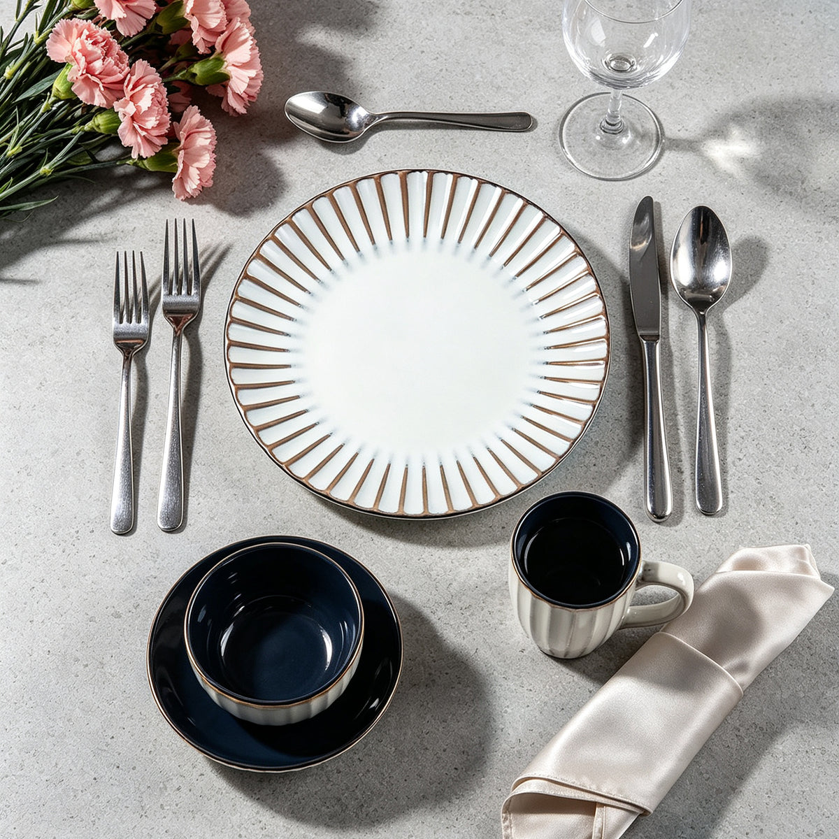 A place setting on a light gray stone surface featuring a white plate with a gold sunburst patterned rim, silver cutlery, a navy blue bowl and mug, pink flowers, and a cream napkin.