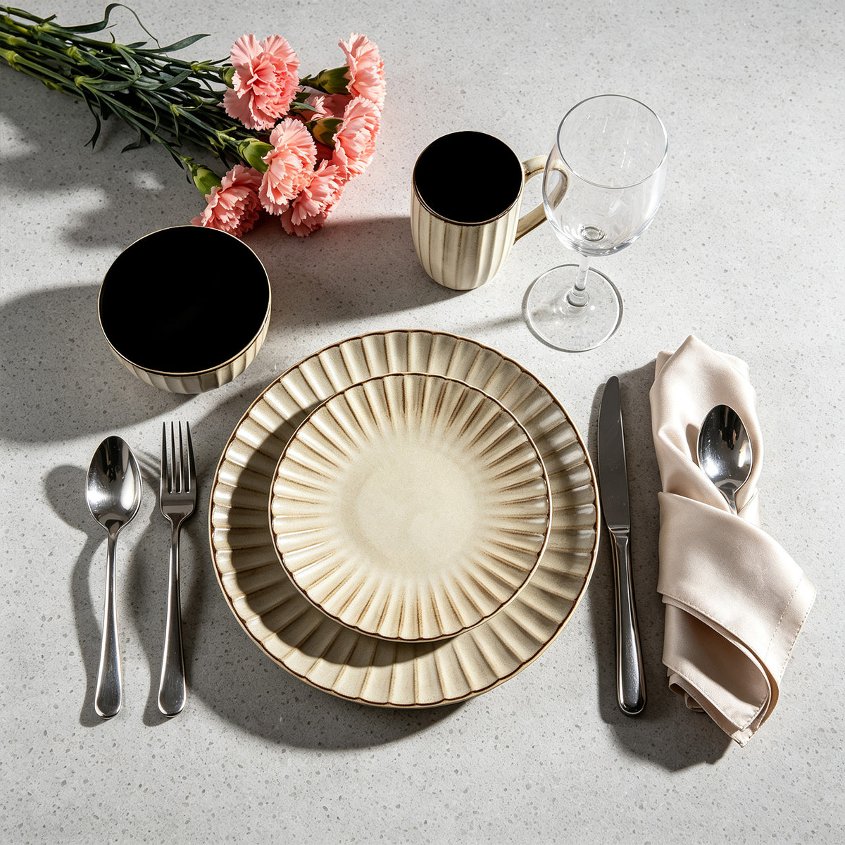 A styled table setting featuring a beige ribbed ceramic dinner plate and matching mug with black interiors, a clear wine glass, silver cutlery, a folded cream napkin, and pink flowers on a light speckled surface.