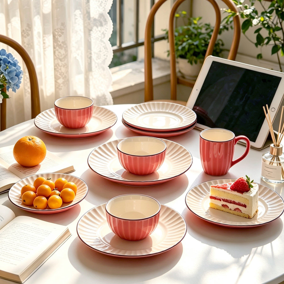 A styled dining table scene with a set of pink and cream ceramic dinnerware, including bowls, mugs, and plates, arranged with food items, a tablet, and a book in a bright, sunlit room.