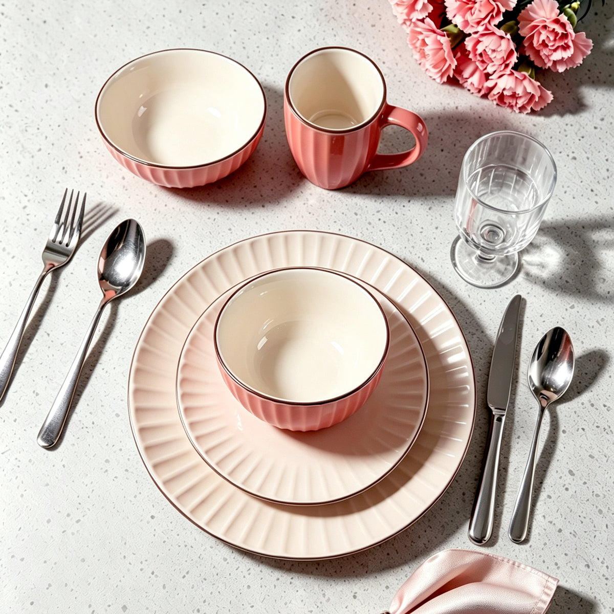 A styled flat-lay of a pink and cream ceramic dinnerware set on a speckled light gray countertop, including a mug, two bowls, a glass, silver cutlery, and pink flowers in the corner.